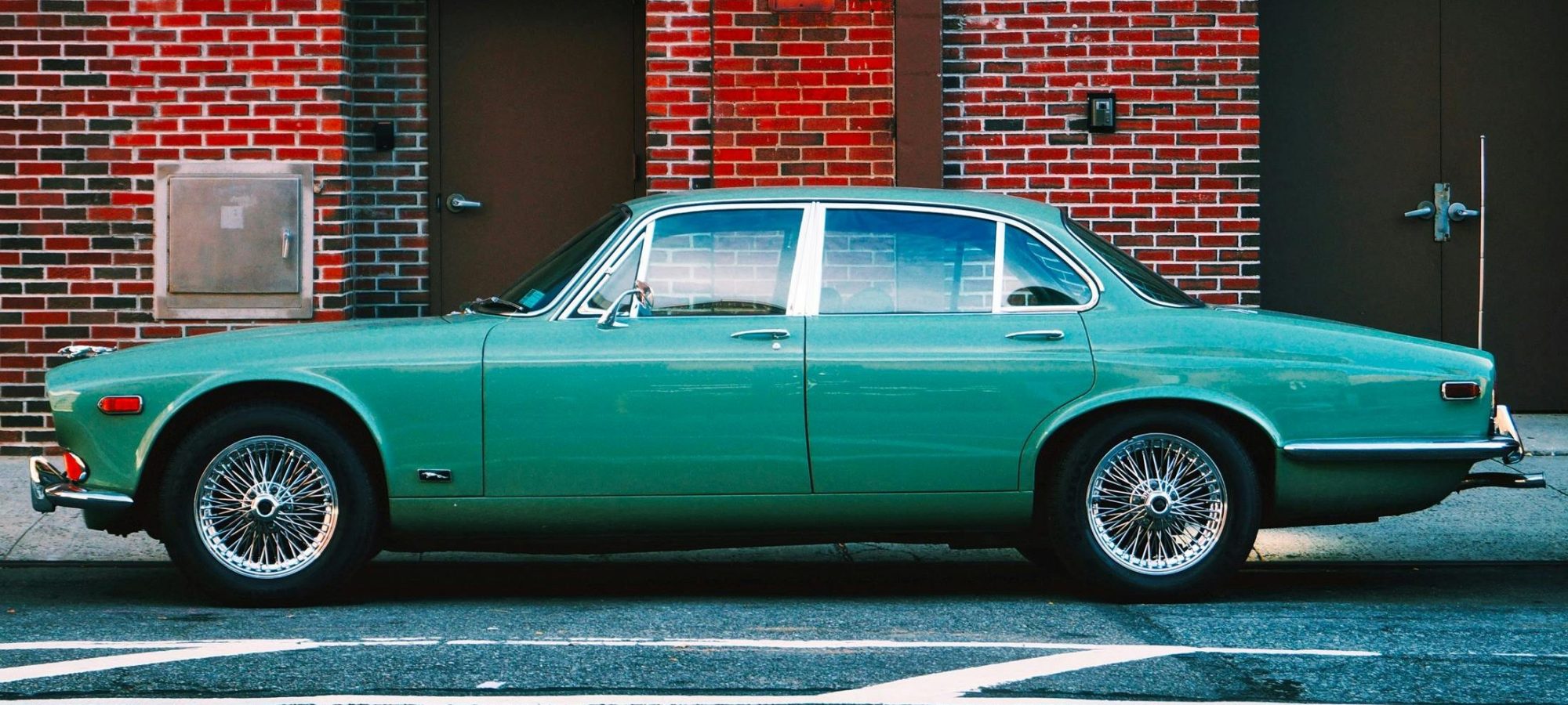 Vintage green Jaguar car parked in an urban setting against a red brick wall, highlighting classic automotive design.