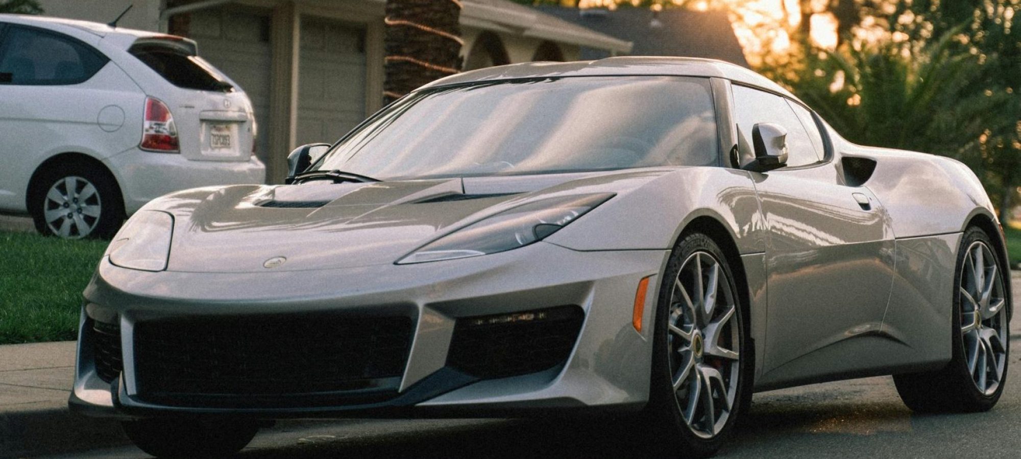 A sleek silver Lotus Evora parked under a palm tree with a sunset backdrop, capturing elegance and leisure.